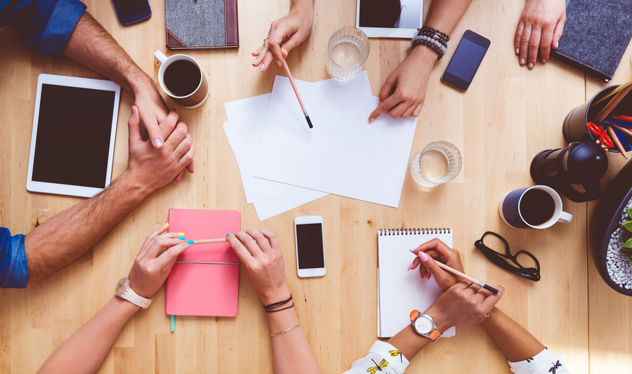 High angle view of group of people sitting at the conference table, discussing, brainstorming. Digital tablets, smart phones, notebooks, coffees on the table. Unrecognizable people.