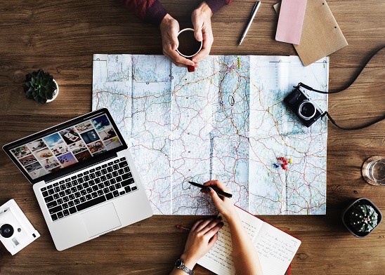Stock photograph of a map sitting in the middle of a table, surrounded by a laptop, a camera, two small pot plants and a notebook. One person's hand, holding a pen, is poised over the map. Another pair of hands cups a coffee mug across from them.